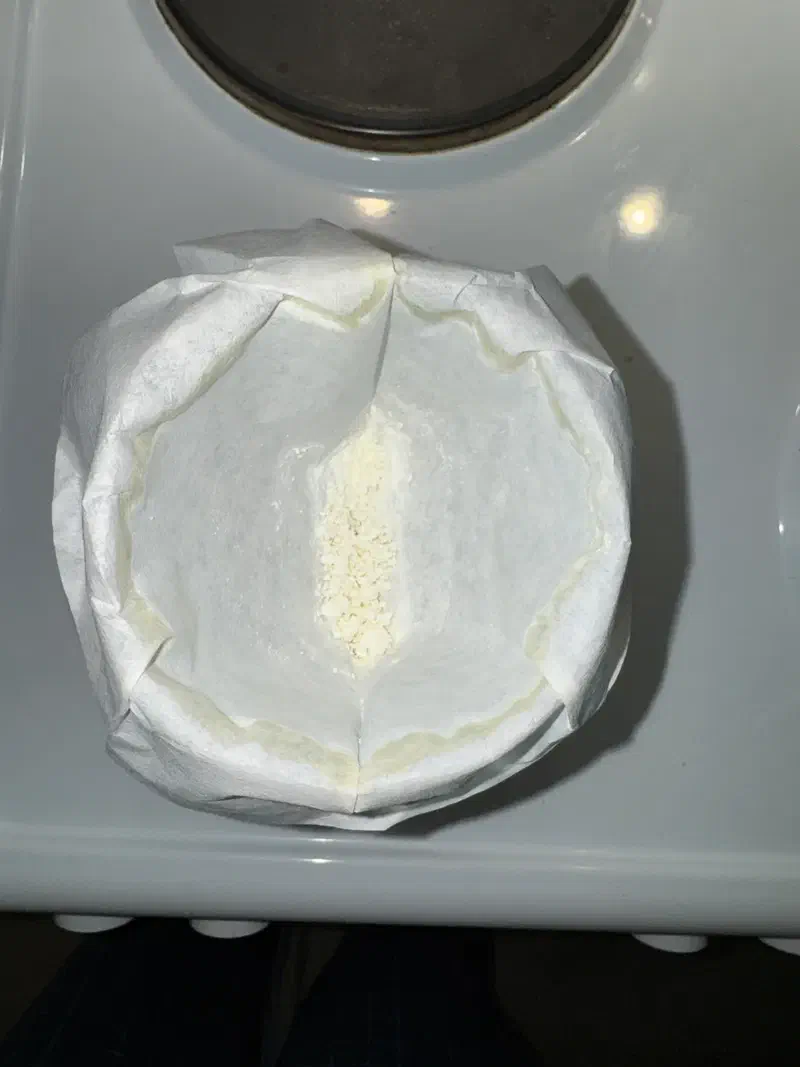 A coffee filter seated in a funnel, seen from above. A pronounced yellow-cream stain rings the entire outer edge of the paper where the solvent front stopped. A small amount of pale yellow powder sits in the centre.