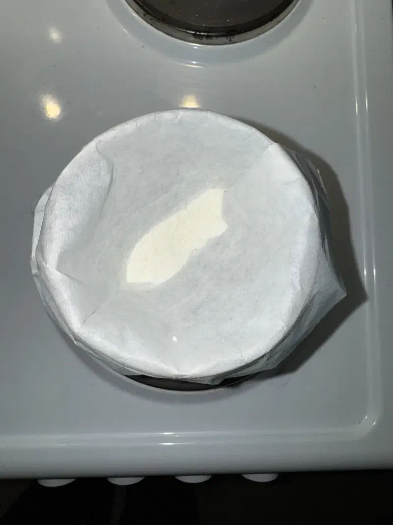 The same filter setup after a third wash. The paper is uniformly white with no coloured ring at the edge. A patch of pale off-white powder sits in the centre.