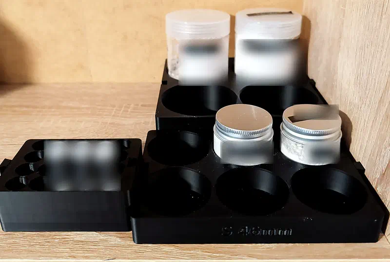 Three black 3D printed sample racks on an oak shelf, holding labelled jars, tins and vials. The labels are deliberately blurred.